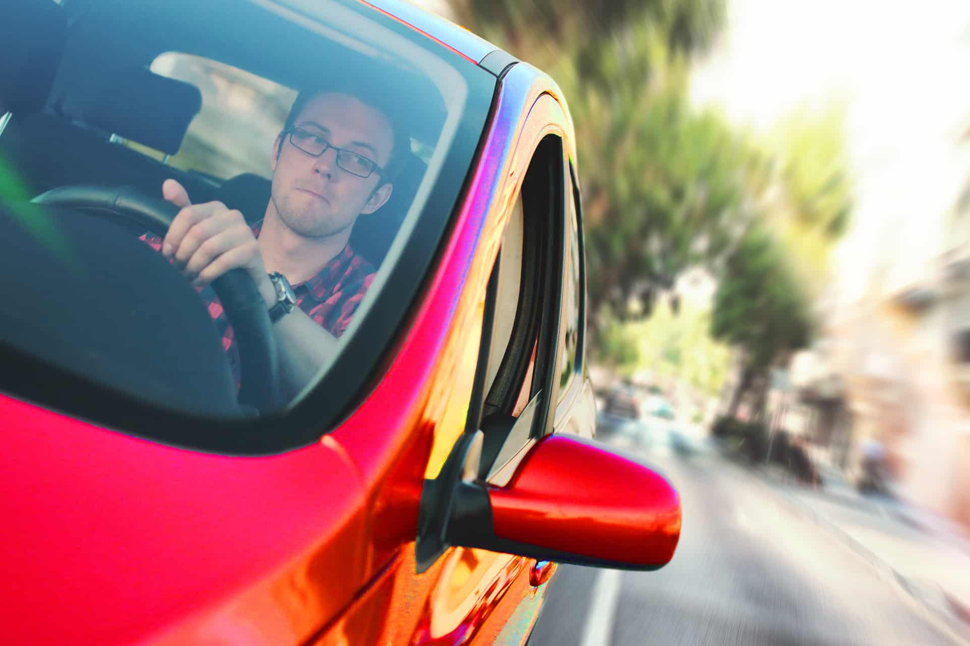 Guy sitting in a vivid red car, seeming to be driving quickly along a road.